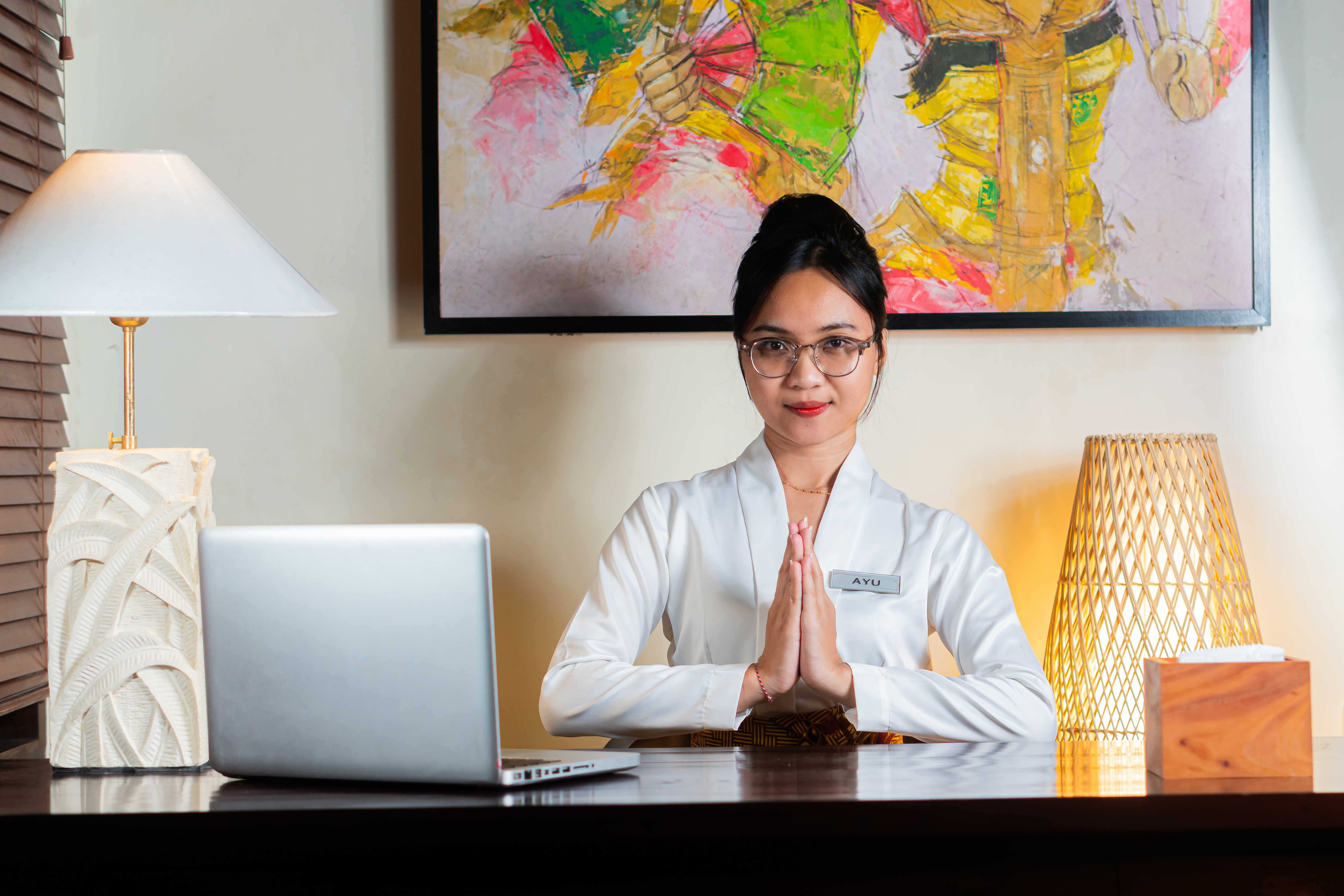 A woman in a white blouse sits at a desk with a laptop, smiling and gesturing with hands together. The setting is a warmly lit room with a lamp and abstract art on the wall.